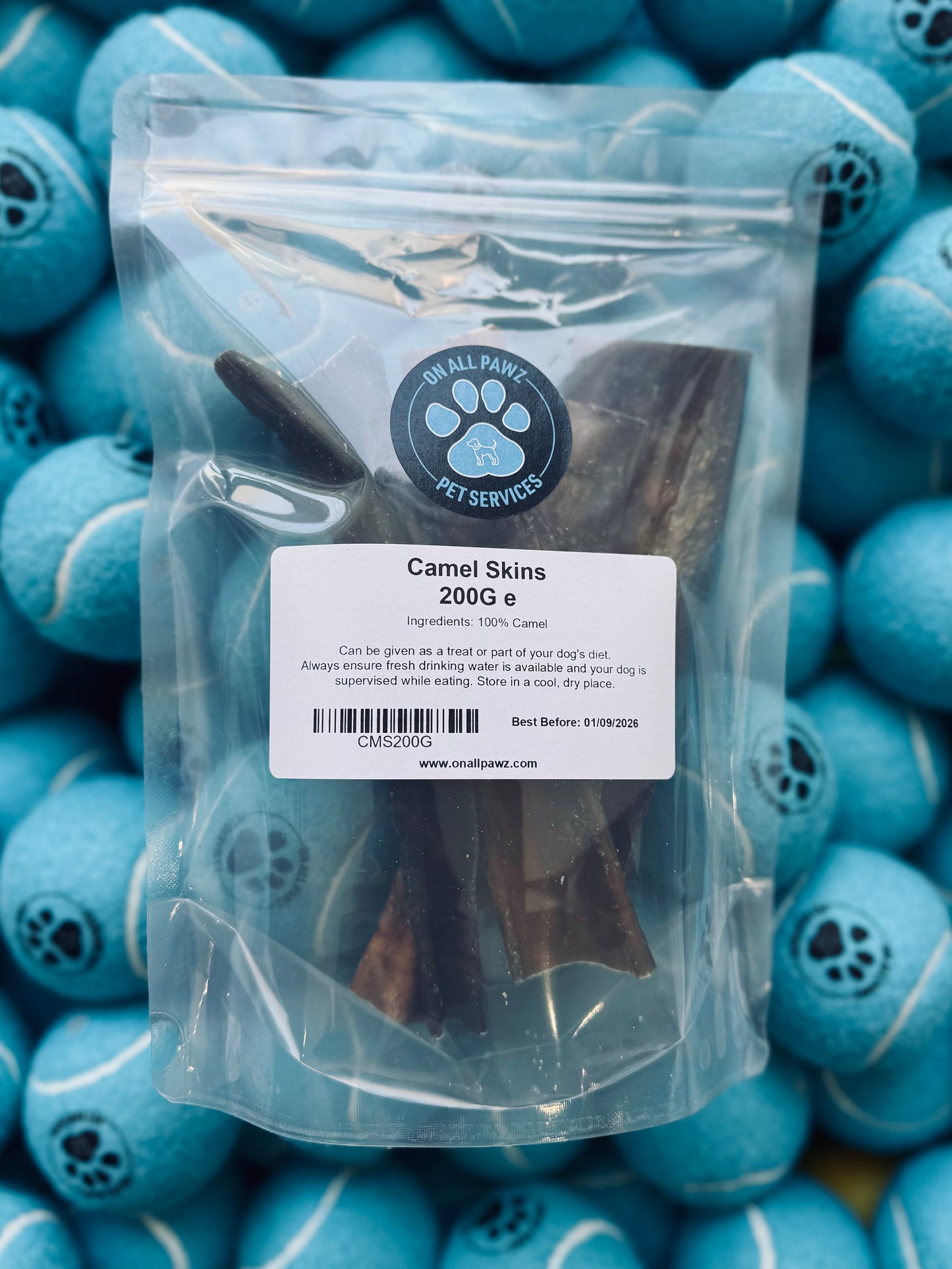 A resealable plastic bag filled with camel skin dog treats, labeled as 200g, with blue dog toys in the background.