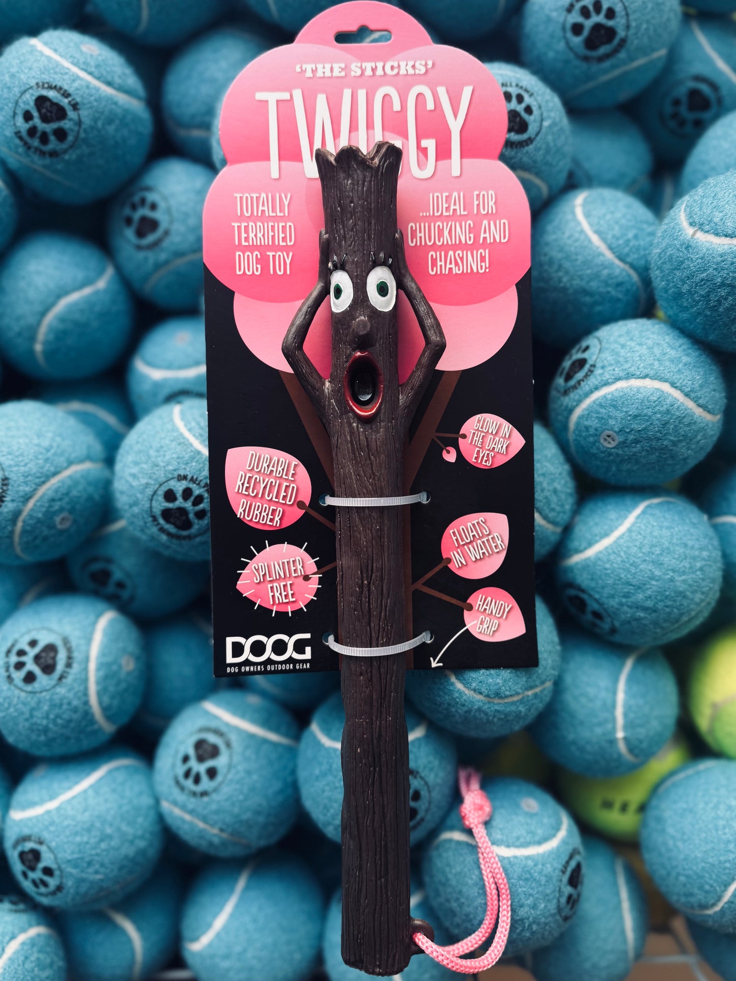 A black DOOG stick dog toy with a cartoonish face and a pink rope attached, presented against a background of tennis balls.