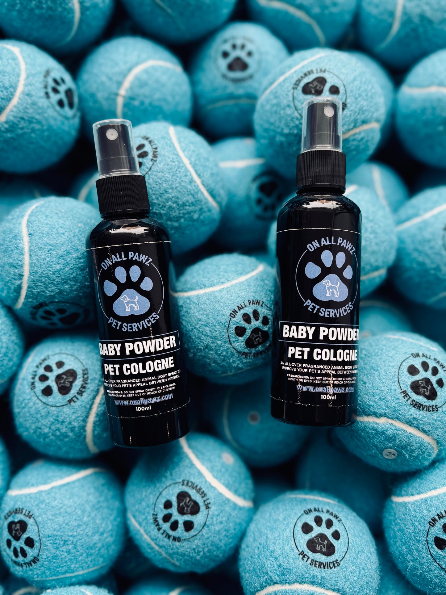 Two bottles of Baby Powder Pet Cologne placed on top of multiple blue tennis balls, with the product's branding visible on the bottles.
