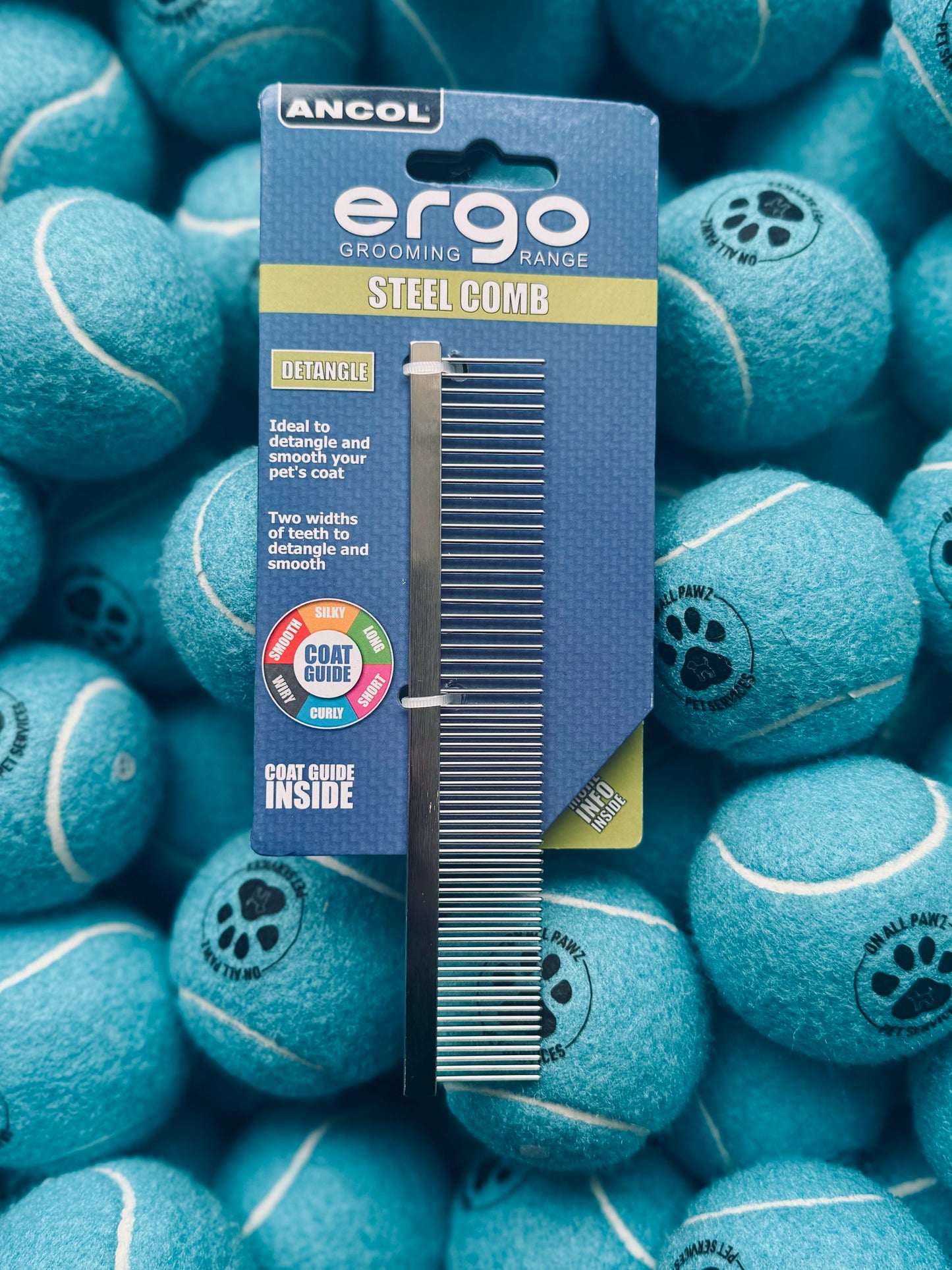 A stainless steel pet comb with dual-length teeth, presented in packaging that reads 'ergo STEEL COMB'. The comb is displayed against a background of blue tennis balls.