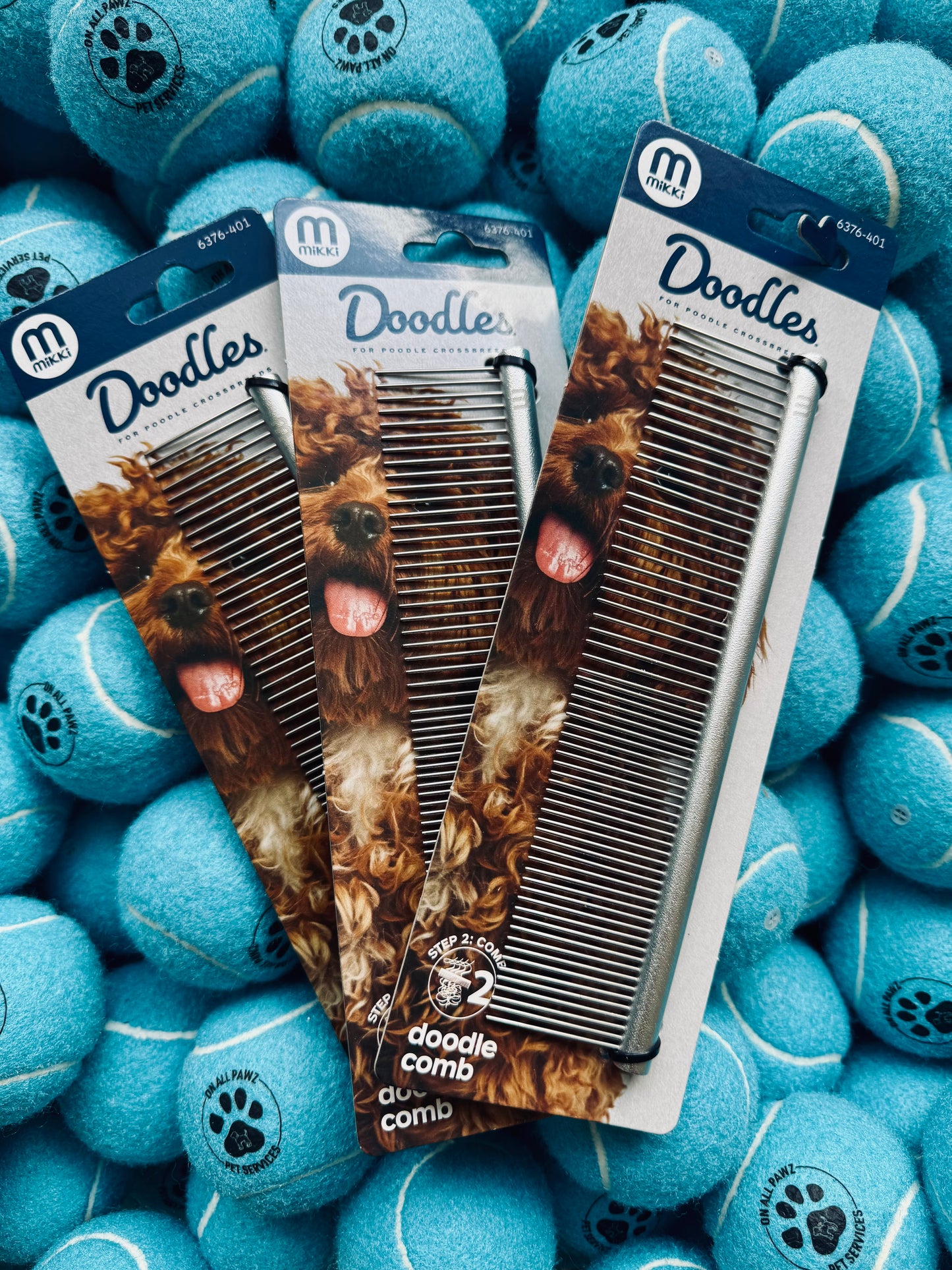 Three dog grooming combs in packaging surrounded by blue tennis balls with paw prints.