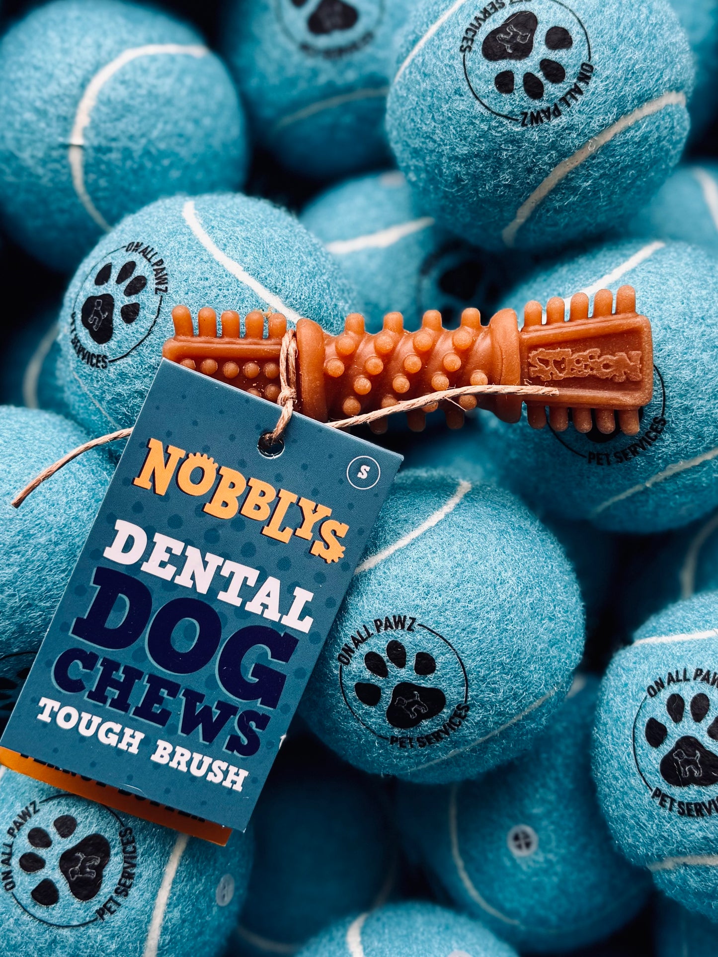 A small blue dog chew toy shaped like a bone with 'NOBBLYS DENTAL DOG CHEWS TOUGH BRUSH' text on it, surrounded by other similar toys.