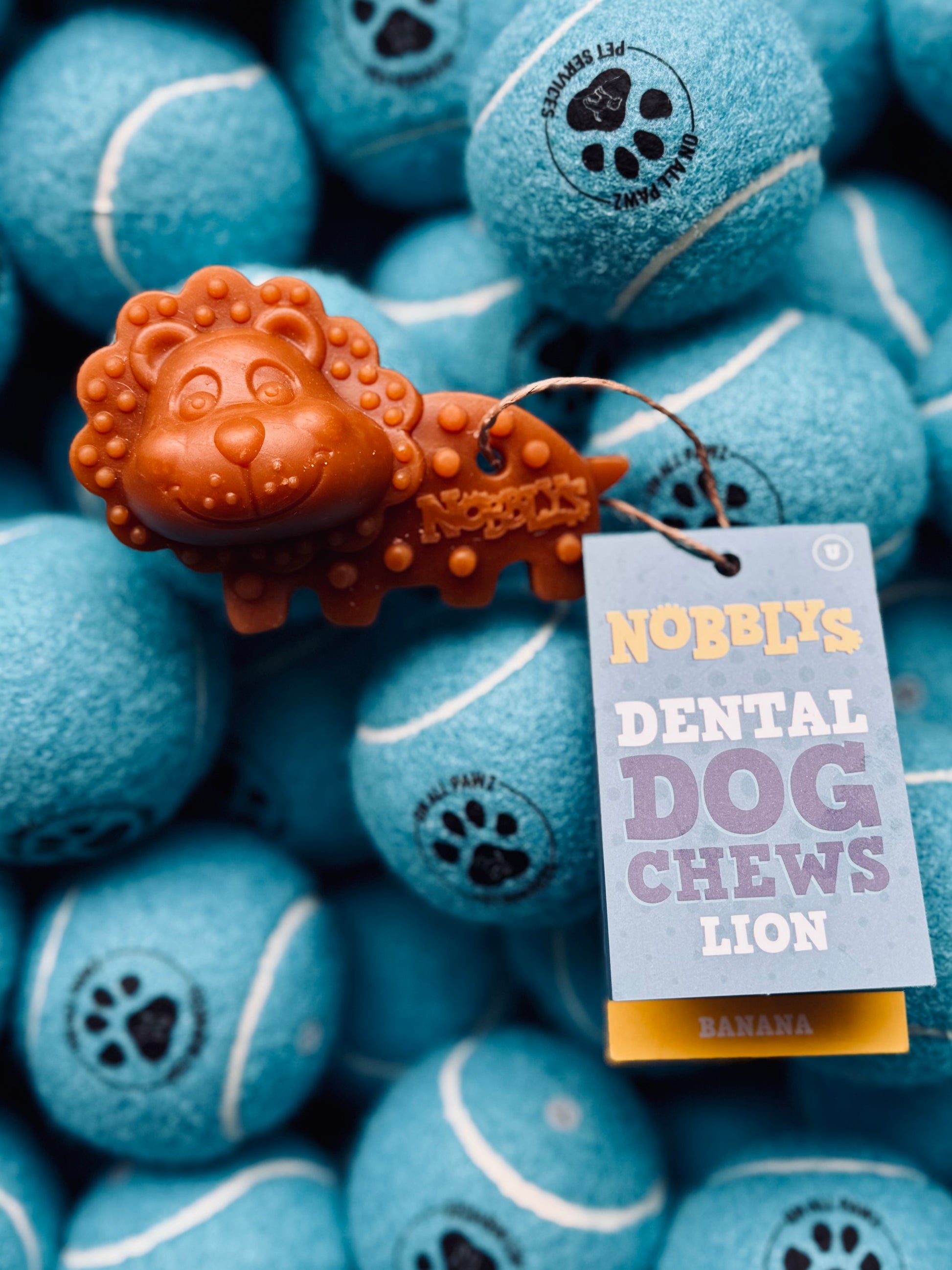 A lion-shaped dental chew treat for dogs, with a banana flavor, placed on a background of blue tennis balls.