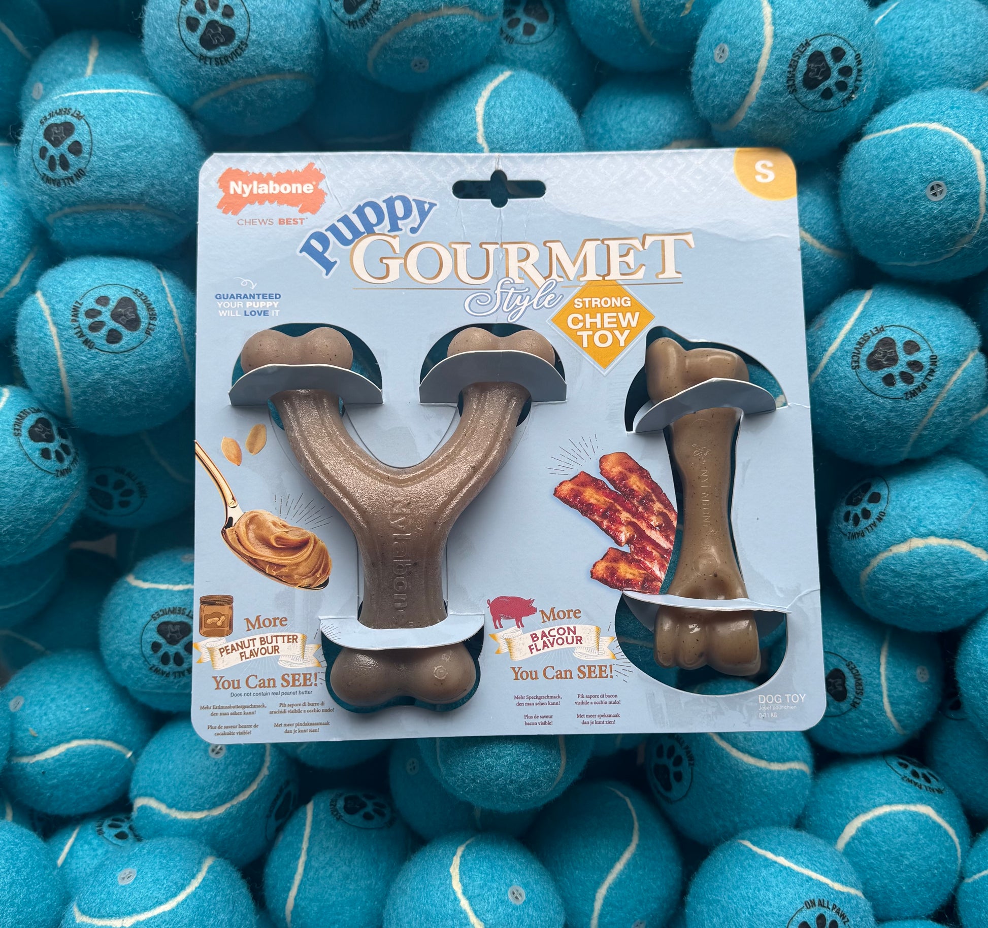 Nylabone Puppy Gourmet Style chew toy packaging with two brown bones on a white background, surrounded by blue tennis balls.