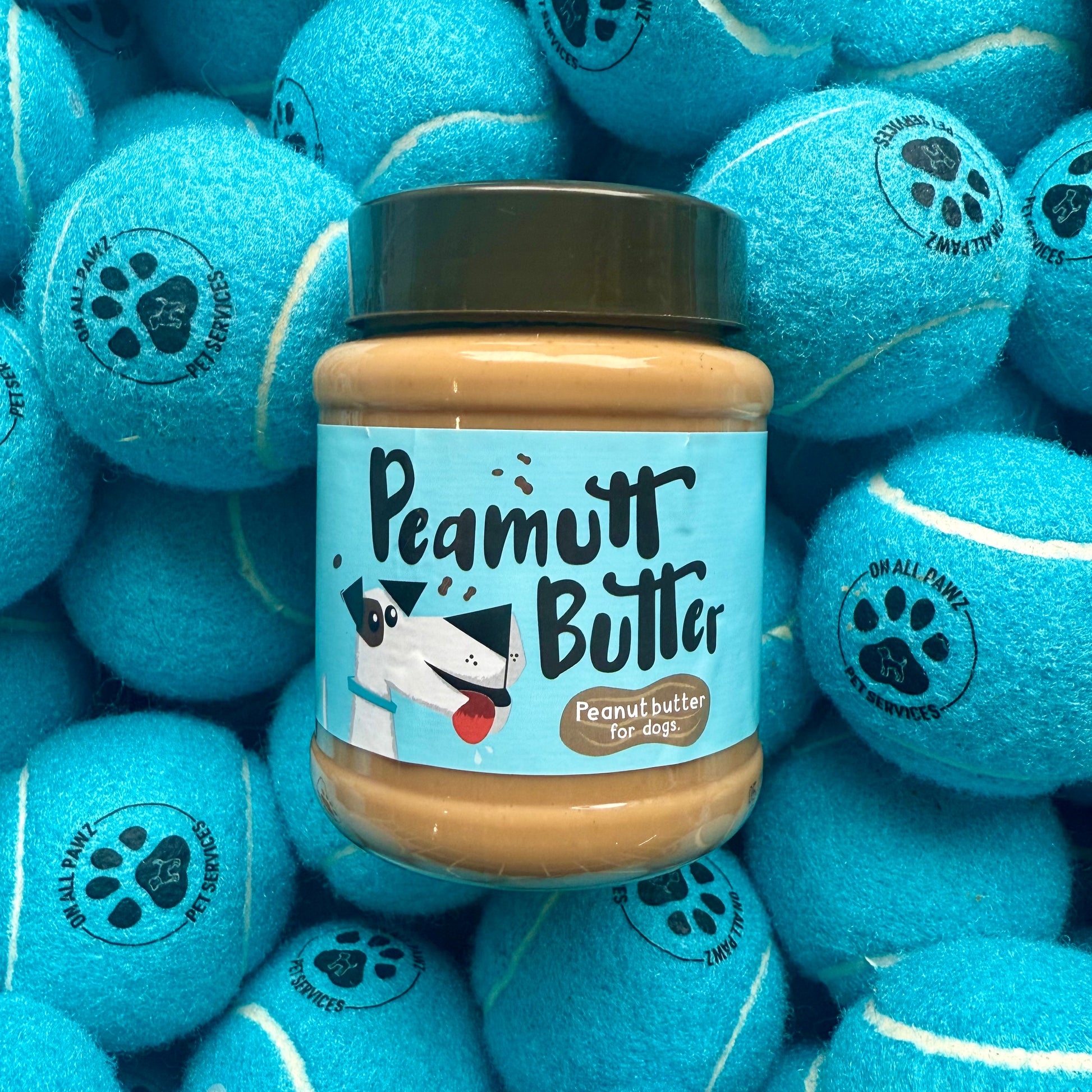 Jar of Peamutt Butter surrounded by blue tennis balls with paw prints on a white background