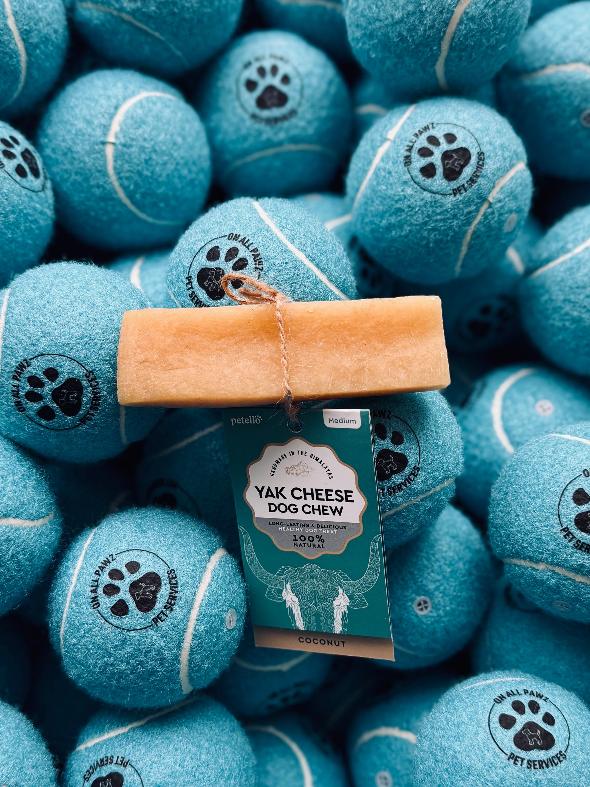 A package of Petello Yak Cheese Dog Chew in Coconut Flavor placed on top of multiple tennis balls, each embossed with a paw print.