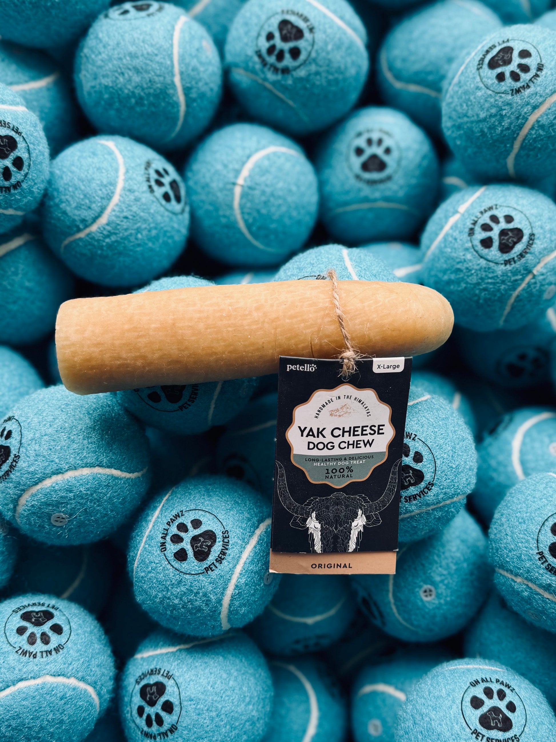 A yak cheese dog chew treat is displayed on a background of blue tennis balls, each imprinted with a paw and bone design.
