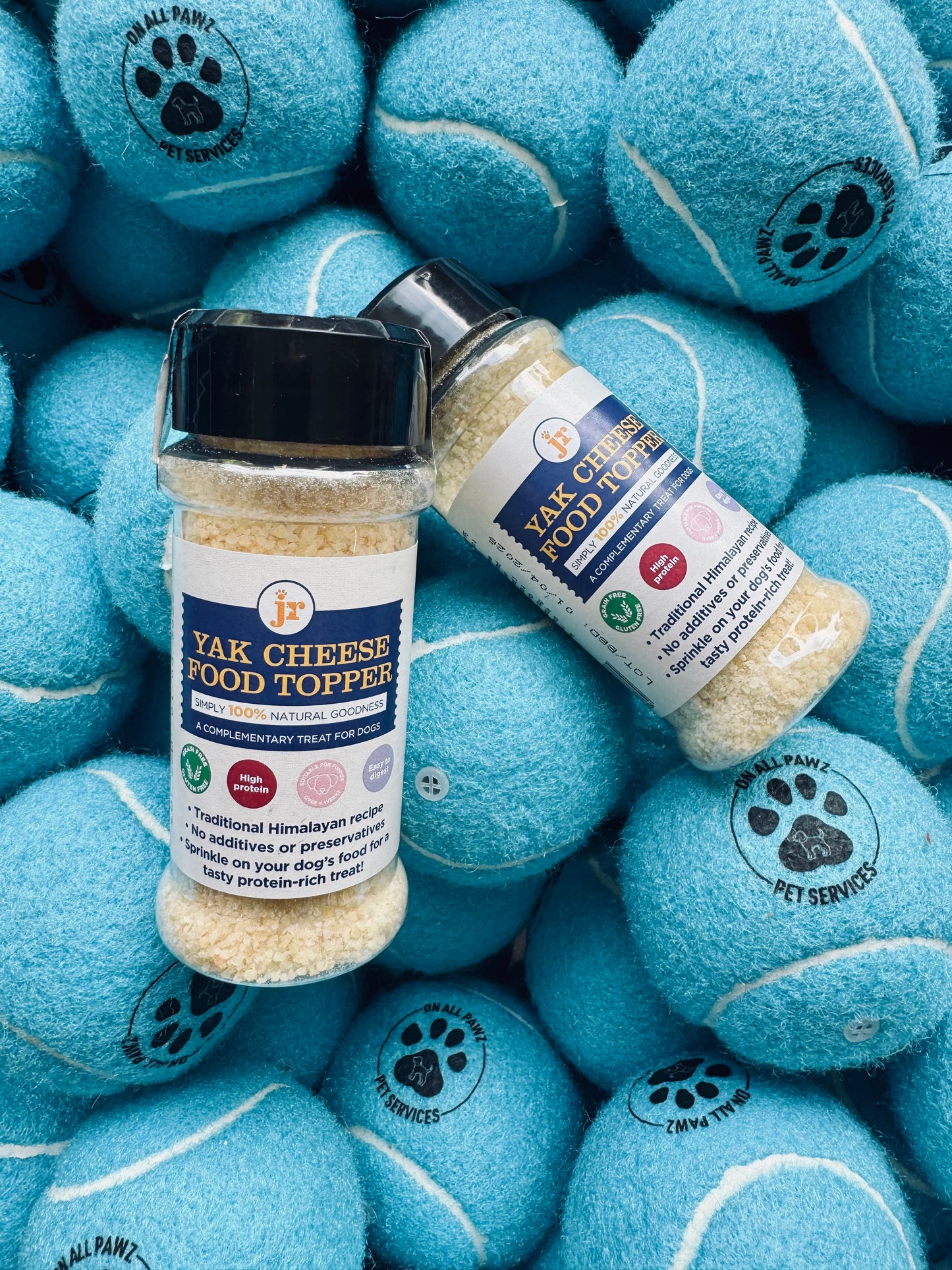 Two bottles of Yak Cheese Food Toppers on a pile of blue tennis balls with paw prints.