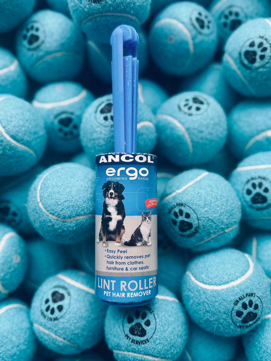 A lint roller with a blue handle and white roller, branded Ancol Ergo, placed on a background of blue tennis balls.