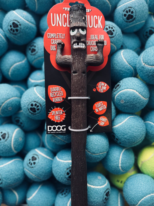 A synthetic dog toy designed to resemble a stick with a cartoonish face, displayed on a tennis ball background. The toy is black with a grinning face and has a rope attached for throwing.