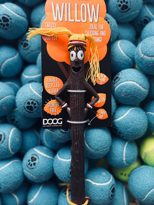 A DOOG Willow Stick dog toy with a cute design, featuring a stick figure with a hat and a built-in rope, displayed on a background of tennis balls.