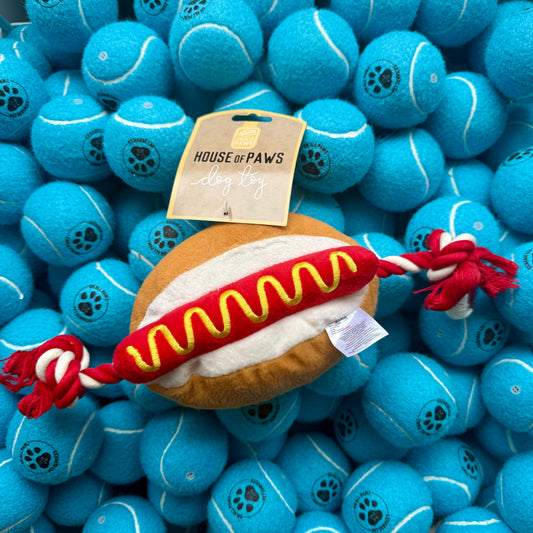 Dog toy shaped like a hot dog bun with a rope on a background of blue tennis balls, branded 'House of Paws'.