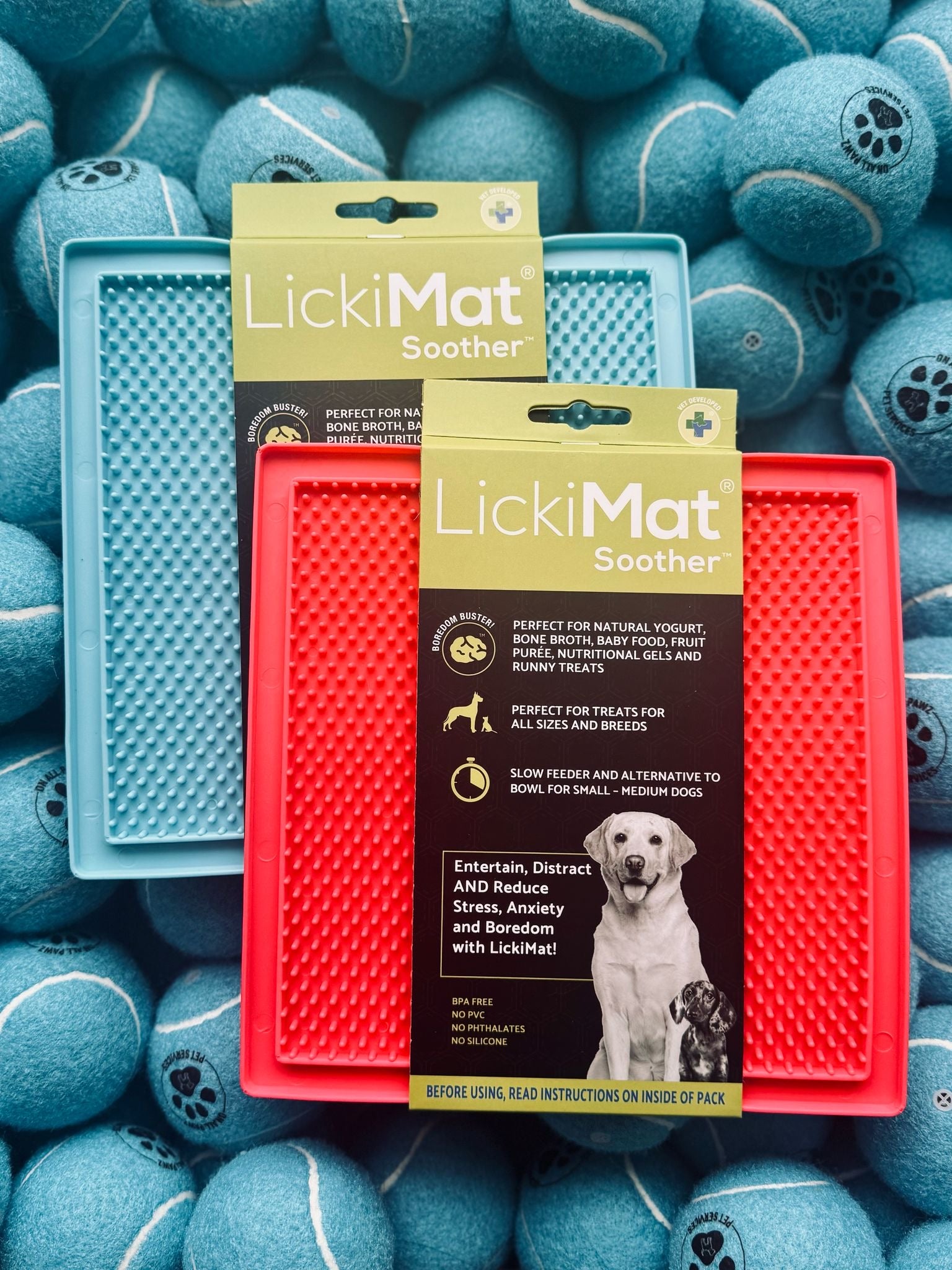 Two LickiMat Soothers, one red and one blue, on a background of blue tennis balls.