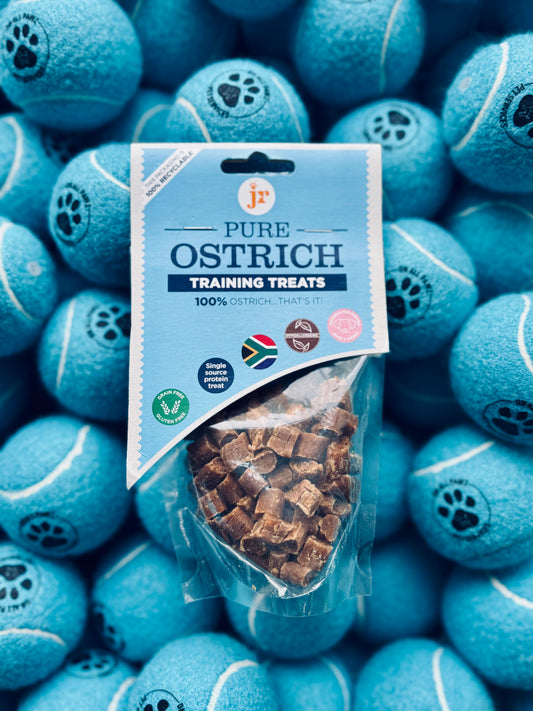 A package of Pure Ostrich Training Treats is displayed in front of a background featuring numerous blue tennis balls. The treat package is transparent, showing the treat pieces inside.