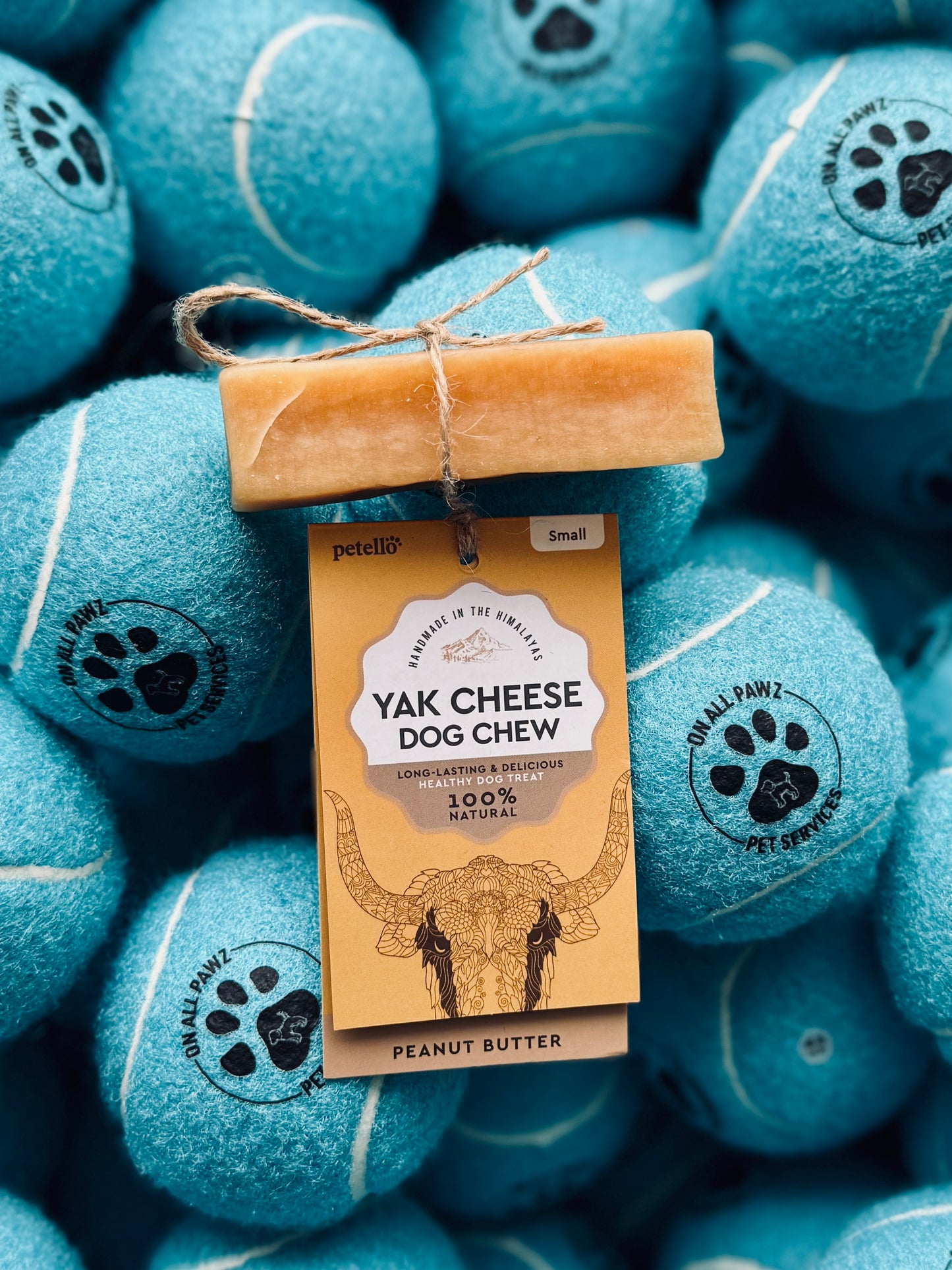 A yak cheese dog chew in peanut butter flavor is displayed on a background of blue tennis balls. The product is packaged with a label that reads '100% Natural Yak Cheese Dog Chew, Peanut Butter Flavor.