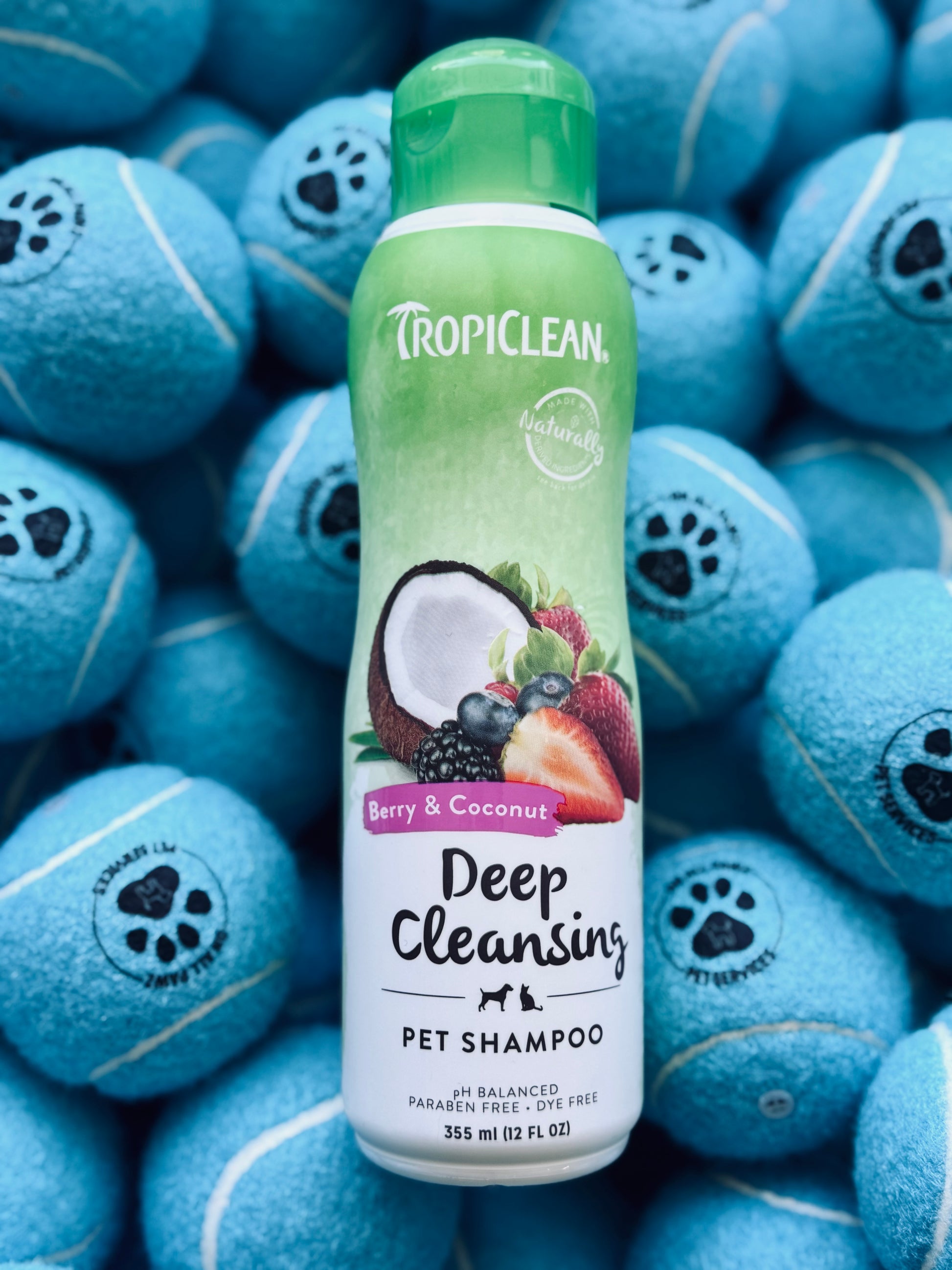 A bottle of Tropiclean Berry & Coconut Deep Cleansing Pet Shampoo, 355ml, with an illustration of a coconut and a strawberry on the label, placed on a background with multiple blue pet toys.
