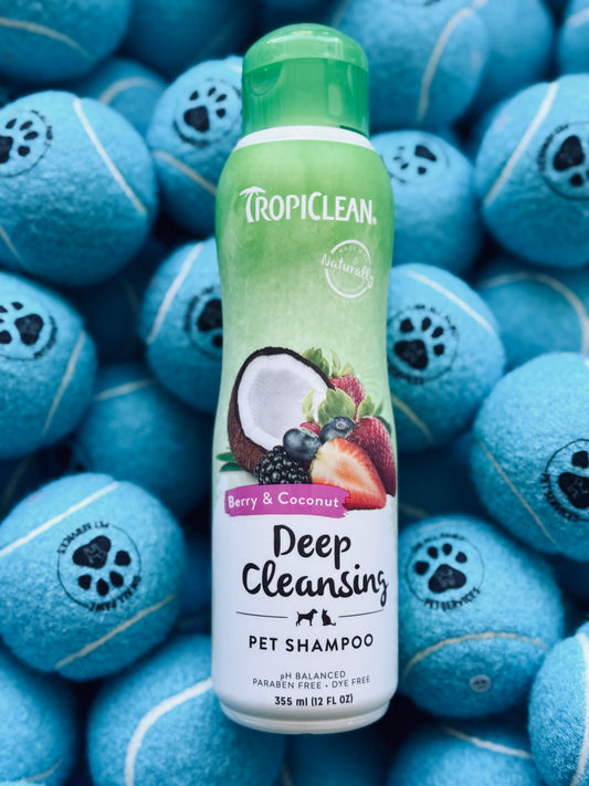 A bottle of Tropiclean Berry & Coconut Deep Cleansing Pet Shampoo, 355ml, with an illustration of a coconut and a strawberry on the label, placed on a background with multiple blue pet toys.